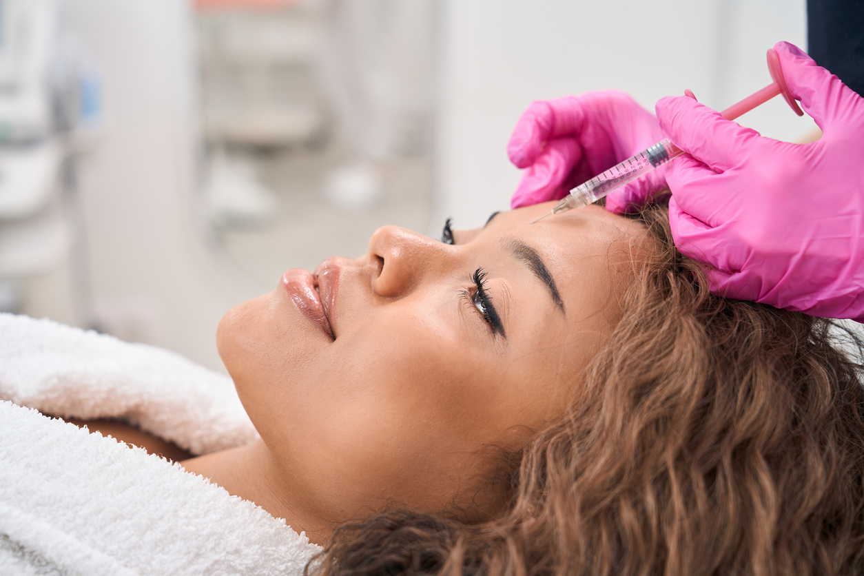 Profile view of a woman lying down with a smile while receiving a non-surgical Botox injection to her forehead from a practitioner wearing pink gloves.