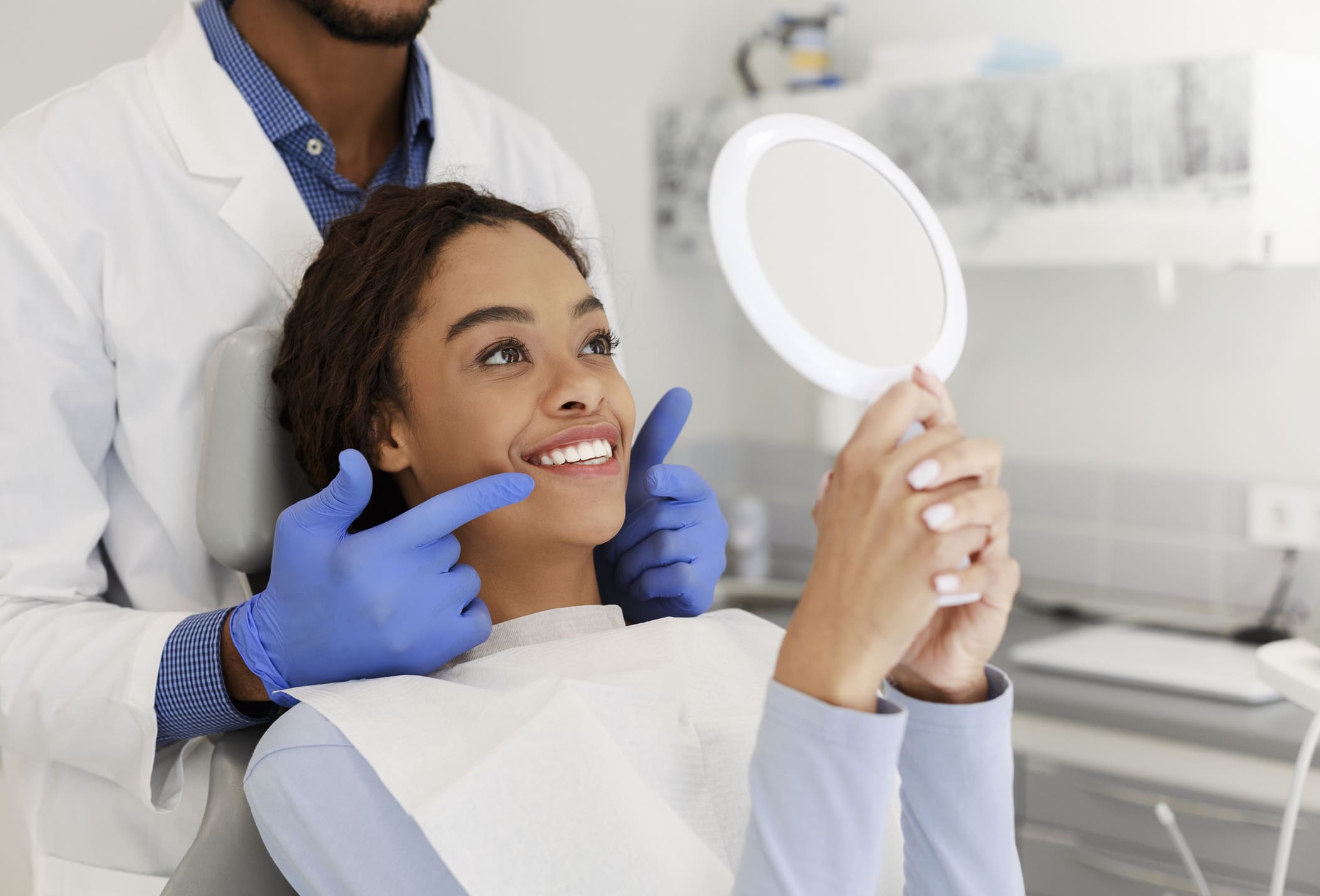 A happy woman sitting in a dental chair looking at her bright white smile in a handheld mirror while a male dentist in a white lab coat and blue gloves points to her teeth.