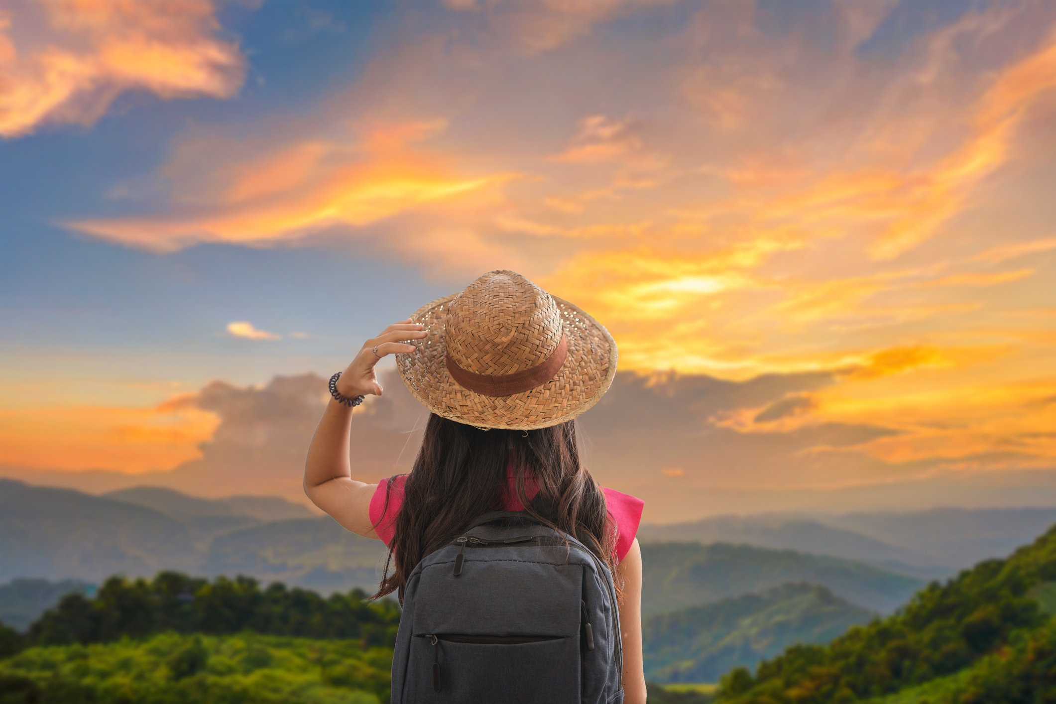 Rear view of a female traveler with a backpack and straw hat looking out over rolling green mountain hills under a vibrant golden sunset sky.