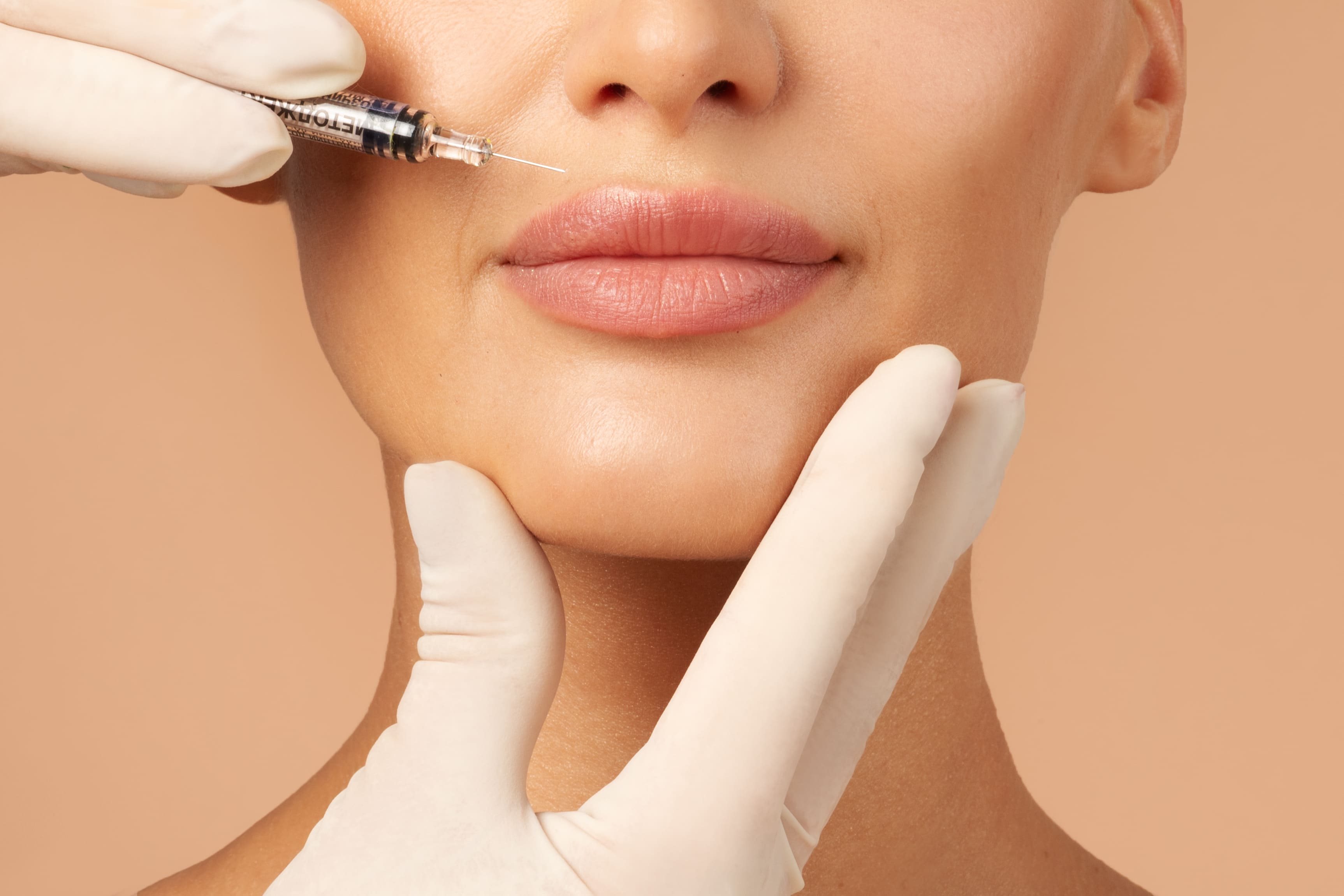 A close-up of a woman receiving a precise cosmetic lip injection from a practitioner wearing white medical gloves.