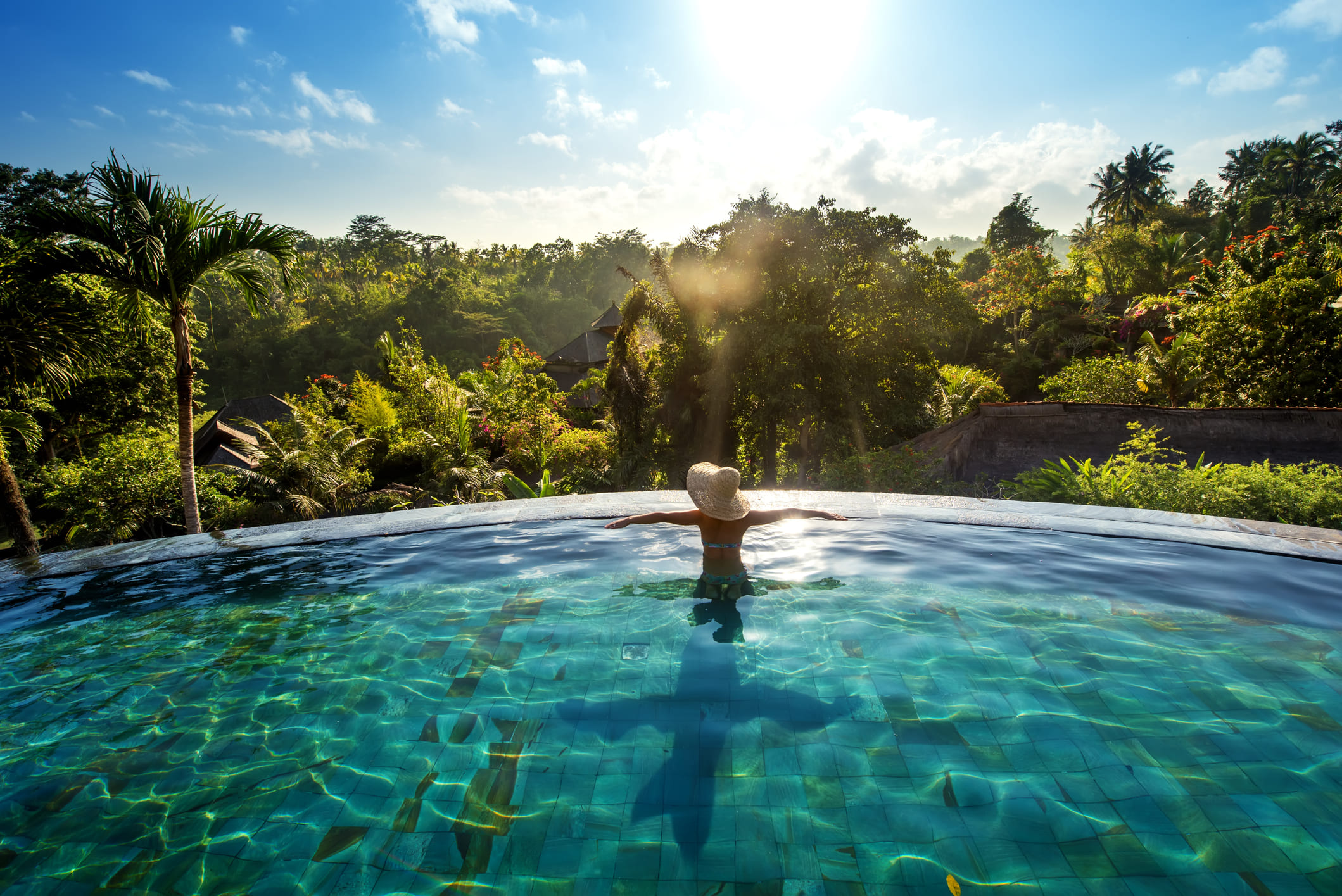 Rear view of a woman wearing a sun hat with her arms outstretched in a luxury infinity pool, overlooking a lush tropical jungle valley bathed in morning sunlight.