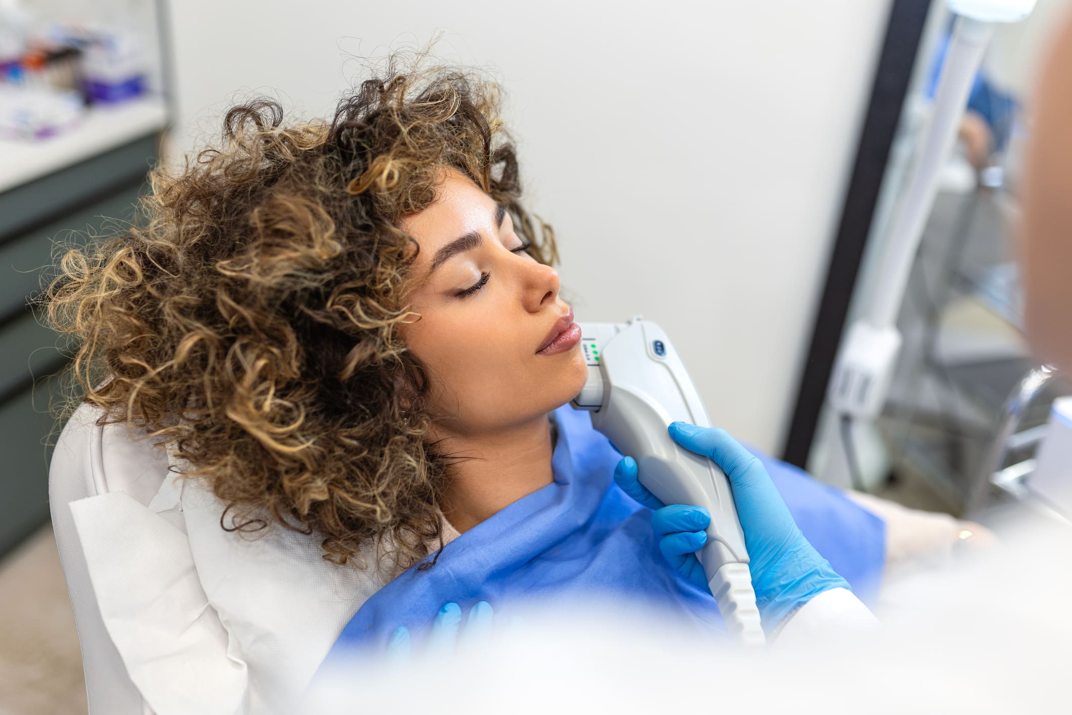A close-up of a woman with curly hair lying back with her eyes closed as a medical professional in blue gloves administers a non-surgical laser skin treatment along her jawline.