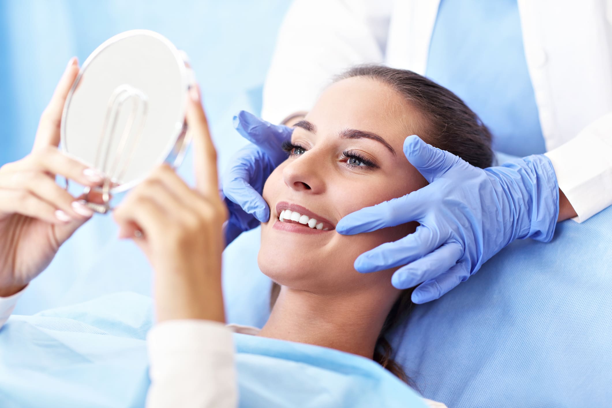 A patient lying back in a clinic chair smiling into a handheld mirror while a professional practitioner in blue gloves frames her face.