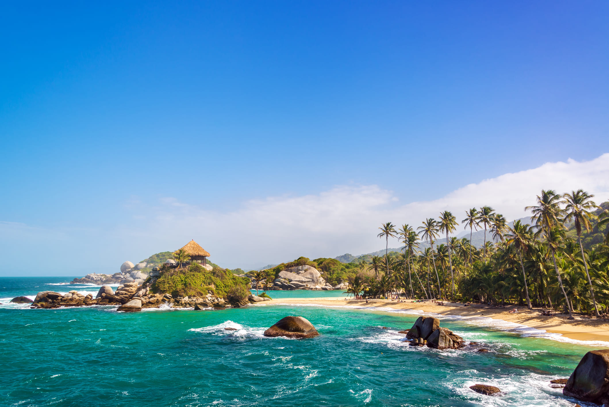 A stunning wide shot of the iconic Cabo San Juan beach in Tayrona National Park, Colombia, featuring a thatched-roof hut on a rocky hill surrounded by turquoise Caribbean waters and a palm-fringed sandy shore under a bright blue sky.