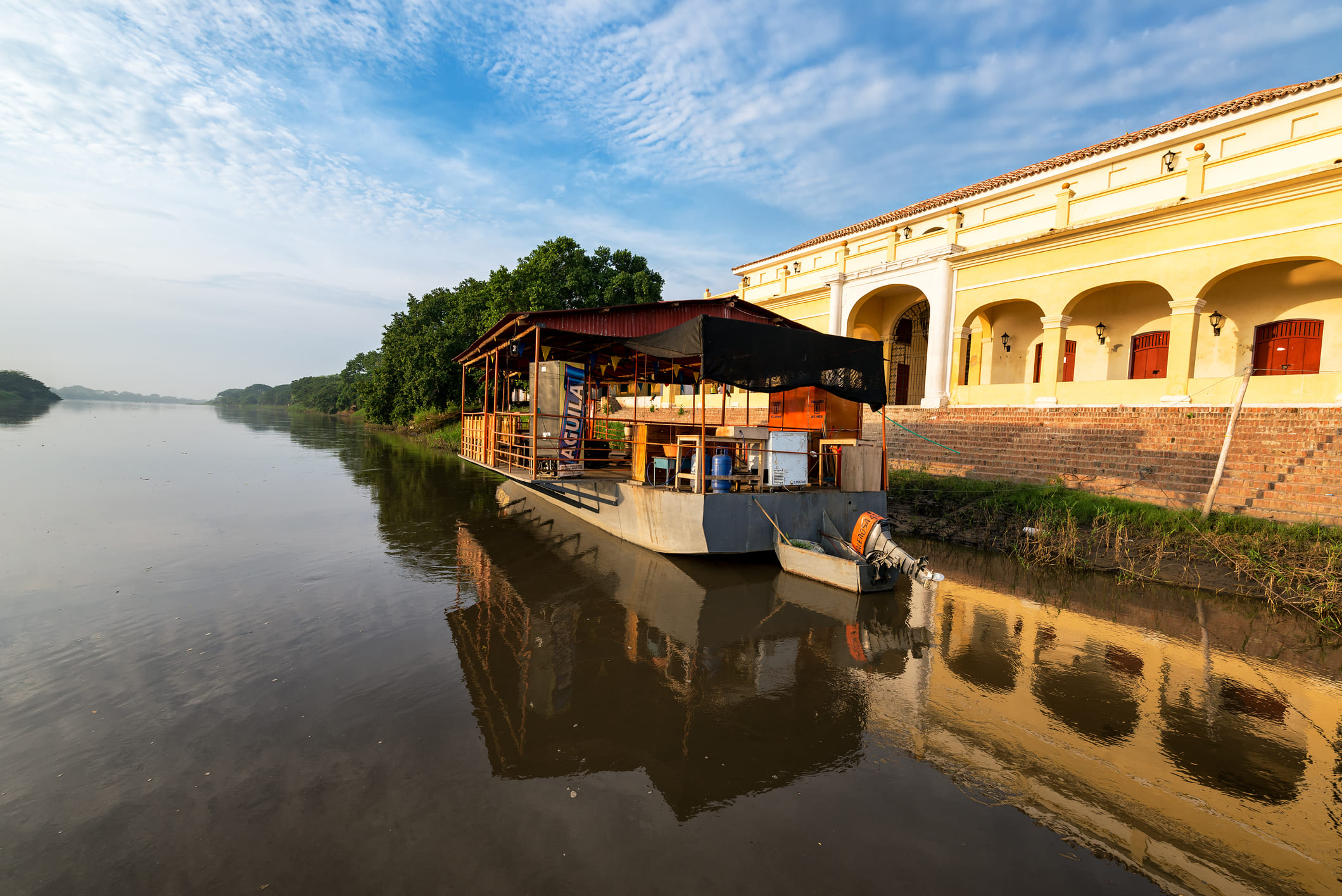 An early morning view of the calm Magdalena River in Mompox, showing a traditional wooden riverboat docked along the water next to a yellow colonial building with grand arches, all perfectly reflected in the still river under a partly cloudy blue sky.