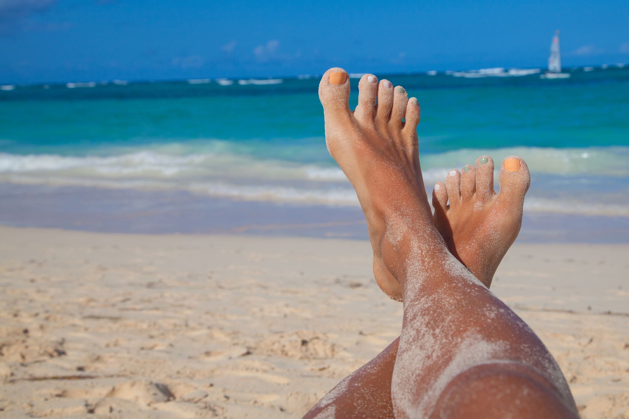 A first-person perspective of a woman's sandy legs and feet crossed while relaxing on a white-sand beach in Tolú, with the turquoise Caribbean Sea and a small sailboat visible under a clear blue sky in the background.